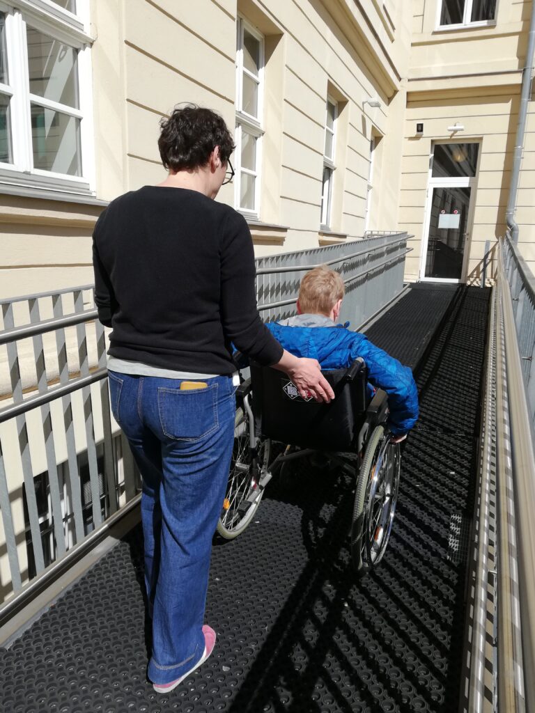 Ein Junge im Rollstuhl versucht die lange Rampe hochzufahren und wird dabei unterstützt.